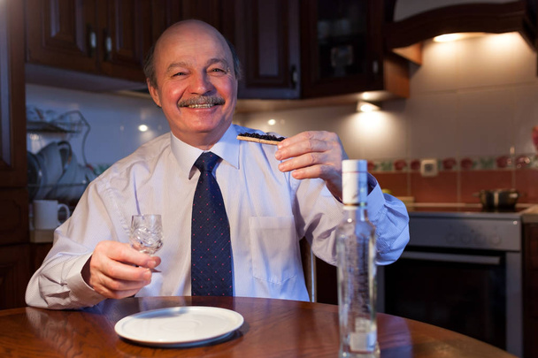 A Russian businessman in a shirt and tie is sitting in the kitchen at the table and pouring himself a pile of vodka.  - Φωτογραφία, εικόνα
