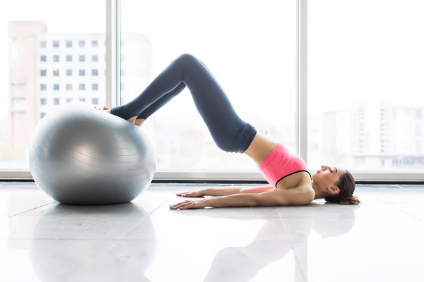Woman and fitness ball. Young woman doing exercises with fit ball at gym. Woman is doing fitness at home on room floor near the window. - Фото, изображение