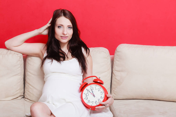 Pregnant woman sitting on sofa holding clock - Fotografie, Obrázek