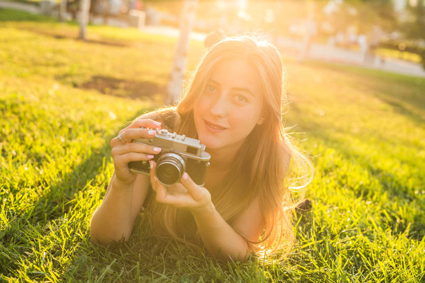 Beautiful girl photographer holds a camera and lying on the grass in the spring outdoors in the park. The concept of tourism and travel. - Photo, Image