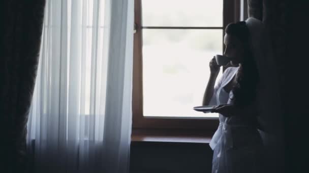Novia joven que se queda cerca de la ventana y toma café por la mañana. Mujer con velo preparándose para la ceremonia de boda
. - Metraje, vídeo