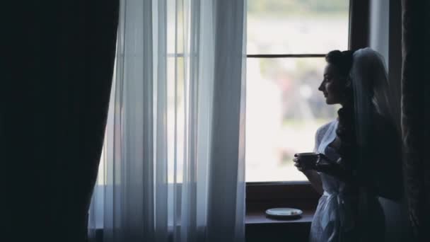 Muotokuva brunette morsian ikkunan lähellä. Nainen valkoisessa elegantissa peignoirissa juo kahvia ja etsii etäisyyttä
. - Materiaali, video