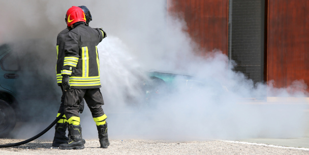 Два пожарных в форме и защитном шлеме тушат огонь.
 - Фото, изображение