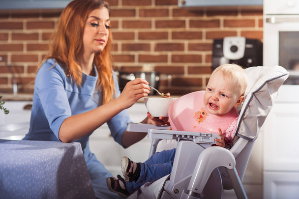 Little baby girl sits in high chair and feeding with spoon her beautiful mother - Photo, Image