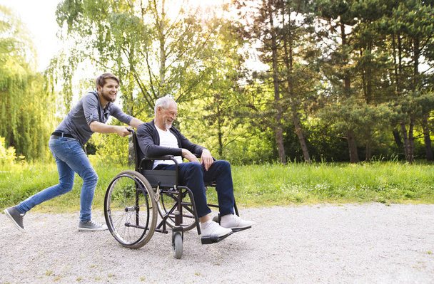 Hipster son running with disabled father in wheelchair at park. - Foto, imagen