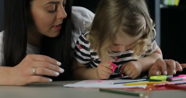 Pretty little girl with blond curly hair and her mother siting on the desk. Girl drawing on a sheet of paper by pink felt pen. Indoor. Close up. - Footage, Video