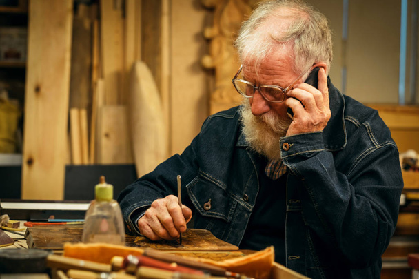 Profissional sênior de escultura em madeira com telefone móvel
 - Foto, Imagem
