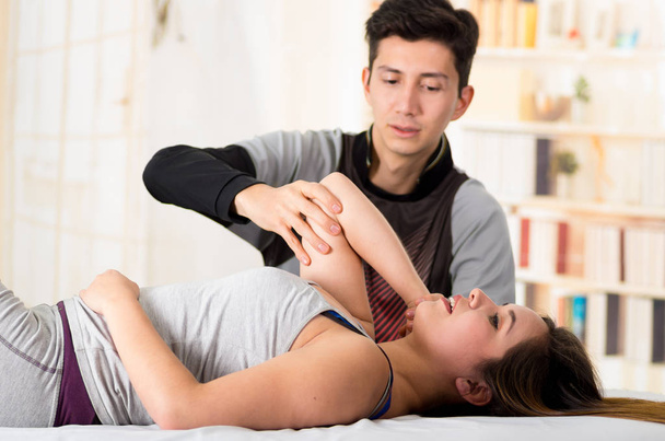 Sports massage. Hnadsome massage therapist massaging elbow of a female athlete, in a doctor office background - Photo, image