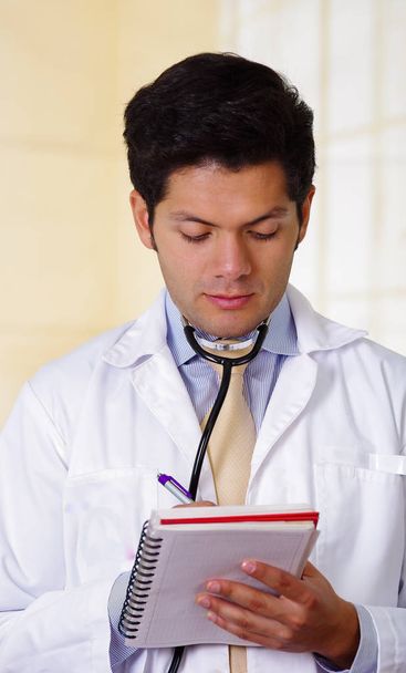 Handsome doctor with an Stethoscope holding from his neck, writing on his notebook notes in office background - Foto, Imagen