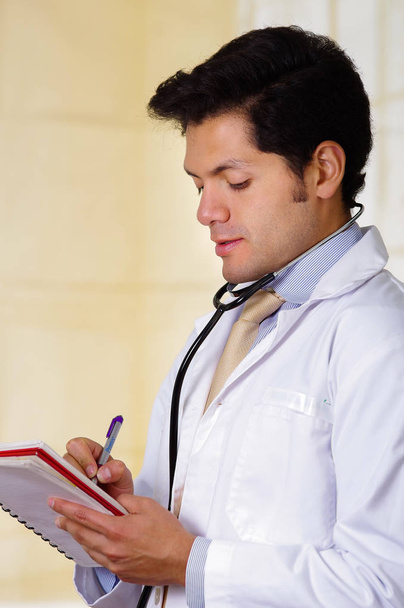 Handsome doctor with an Stethoscope around his neck, writing on his notebook notes in office background - Photo, Image