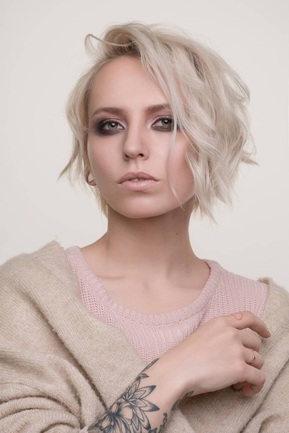 Portrait of girl of blonde with dark eye makeup and short hair in a light pink sweater standing on a light background looking into the camera, the photo below - Photo, image