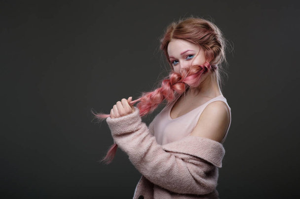 Portrait of girl with pink hair in braids, pink and blue makeup, standing in a half-circle on a dark background playfully looking into the camera and covering the face braid - Foto, immagini