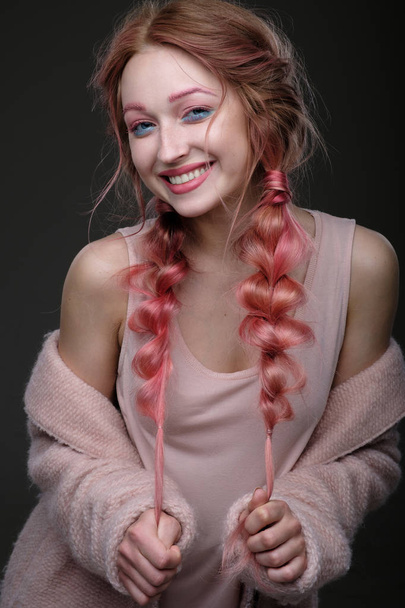 Portrait of a girl with pink hair in braids, and blue and pink makeup, a pink coat, worn off the shoulder, standing and pulls the ends of the braids and looking at the camera, smiling widely - Zdjęcie, obraz