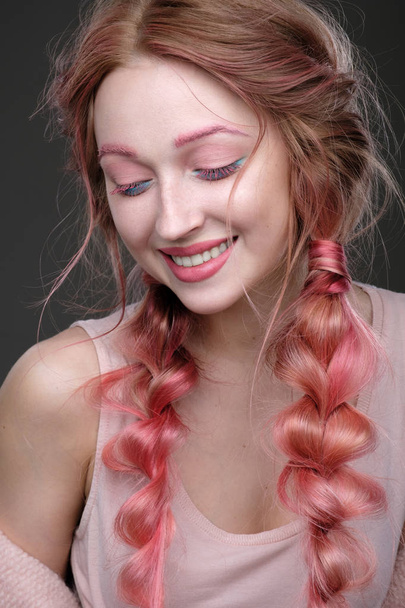 Крупним планом портрет дівчини з рожевим волоссям в коси, рожево-блакитний макіяж. Дівчина стоїть на темному тлі, очі закриті і усміхнені
 - Фото, зображення