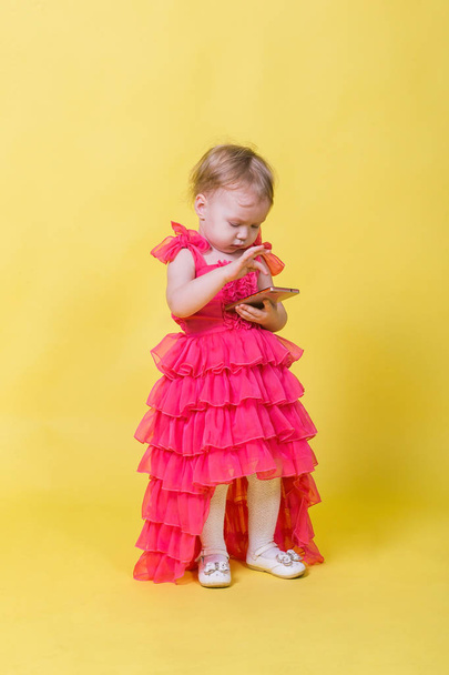 Girl toddler in a pink dress on a yellow background holding a smartphone - Fotografie, Obrázek