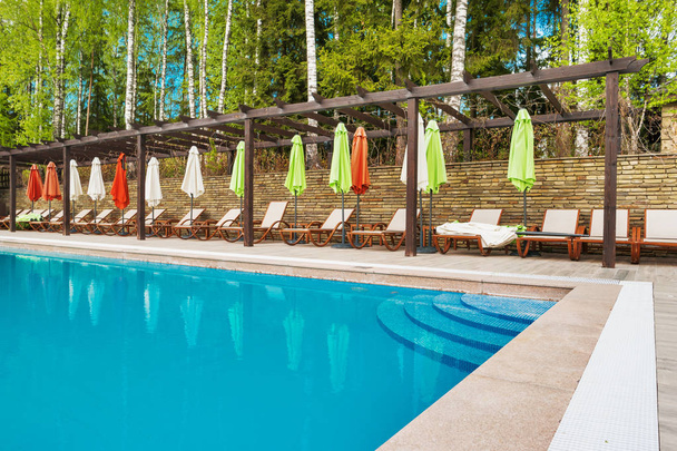 Lehátka a slunečníky u bazénu s jasně modrou vodou. Březový les strom kolem bazénu. - Fotografie, Obrázek