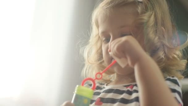 Portrait of side view of pretty caucasian young girl with blond curly hair making bubble blower on living background. Indoor. - Materiał filmowy, wideo