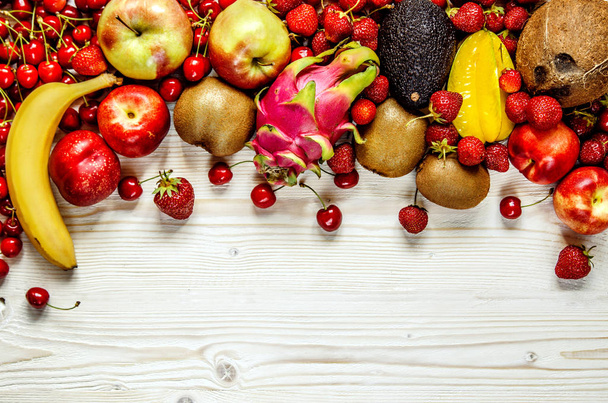 Frutas de cereza, fresa, plátano, aguacate, carom, coco, melocotón, manzana y fruta de dragón yacen sobre una superficie de madera blanca
. - Foto, Imagen