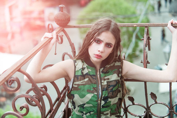 Pretty girl in camouflage vest holding metallic balcony rails - Foto, afbeelding