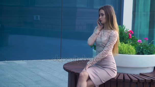 girl sitting on a circular bench near flowers and speaks on a mobile phone - Felvétel, videó