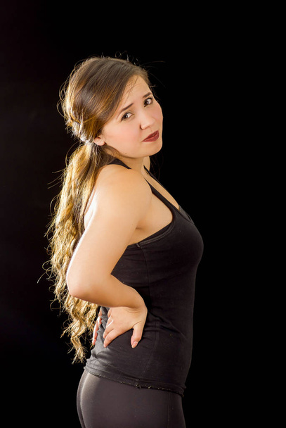 Young beautiful sportwoman suffer cervical pain in her lower back, in black background - Fotografie, Obrázek