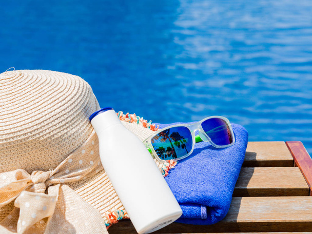 Pláž hat, sluneční brýle, modrý ručník a opalovací krém na okraji bazénu. Dovolená, pláž, letní cestování koncept - Fotografie, Obrázek