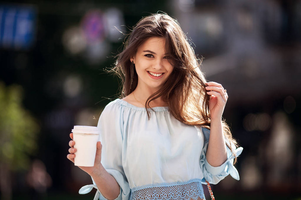Close up fashion street style portrait of a beautiful girl in a casual outfit walks in the city . Beautiful brunette Keeps coffee. - 写真・画像