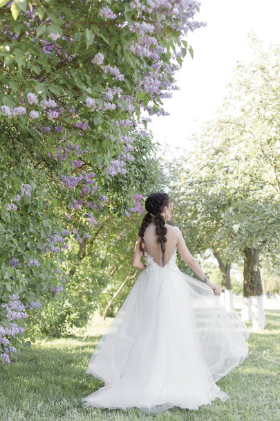 Nabídka krásná roztomilá dívka nevěsta v bílém vzduchu šaty s kytici šeříku v jejích rukou, chůze přes park na slunný jarní den. Foto v jemné barvy - Fotografie, Obrázek