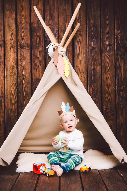 Child, boy, playing in tent indoors on a wooden background. Dressed in Indian - Foto, Bild