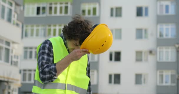 Attractive caucasian builder in a green vest going on unfinished construction background putting on the yellow helmet, looking around. Outdoor. - Metraje, vídeo
