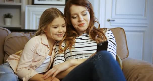 Happy attractive woman with her daughter spending time together at home on the couch. Mother and daughter looking at the smart phone, smiling, hugging and laughing. - Кадри, відео