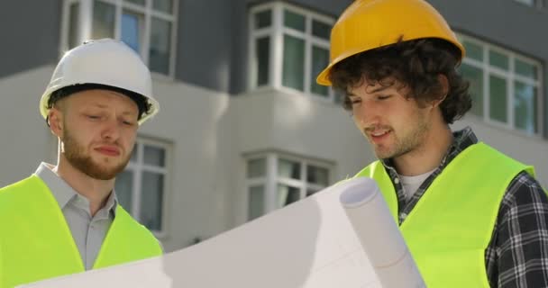 Portrait of two caucasian builders in the helmet and special clothes analysing drawing of building and talking about it. Outdoor. - Séquence, vidéo