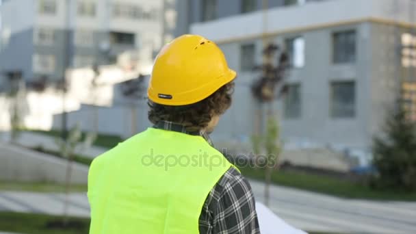 Back view of architect in yellow helmet and green vest standing on building background, analysing drawing. Outdoor. - Séquence, vidéo