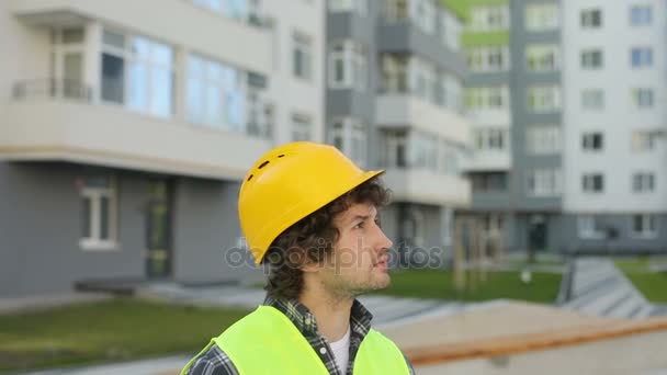 Portrait of caucasian builder in yellow helmet and green vest looking drawing standing on unfinished construction background. Outdoor. - Séquence, vidéo