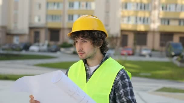 Concentrated caucasian builder in yellow helmet and green vest analysing drawing standing on unfinished construction background. Outdoor. - Video