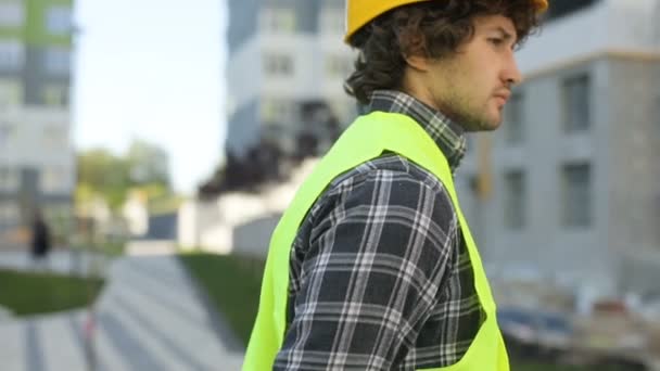 Side view of serious caucasian builder in yellow helmet and green vest on unfinished construction background. Close up. - Záběry, video