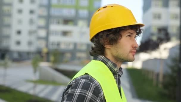 Portrait of side view of concentrated caucasian builder in yellow helmet and green vest standing on unfinished construction background. - 映像、動画