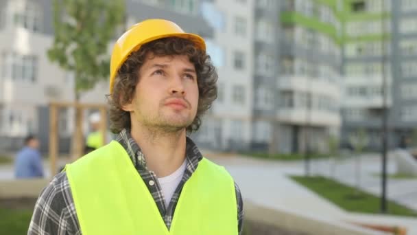 Portrait of builder in yellow helmet and green vest standing on unfinished construction territory, looking up. Outdoor. - Video