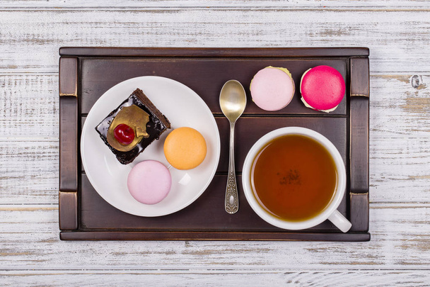 Cup of tea, piece chocolate cake and macaron cakes on tray on white wooden table. Lifestyle concept. - Foto, imagen