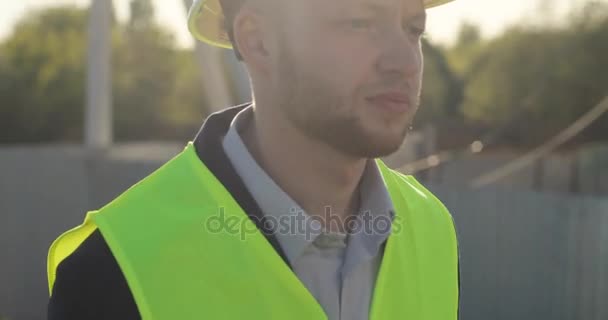 Portrait of serious caucasian builder in a green vest going at initial stage of construction. Outdoor. - Imágenes, Vídeo