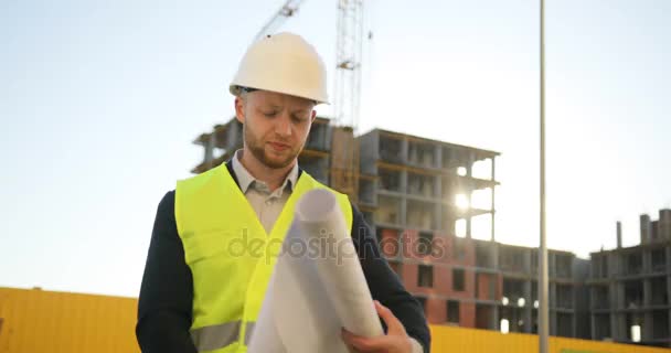 Young architect in white helmet standing on building background. Caucasian engineer looking drawing. Outdoor. - Video, Çekim