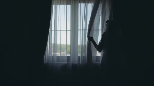 Silhouette di giovane sposa in peignoir bianco che rimane vicino alla finestra. Donna guarda la distanza, mattina prima del matrimonio
. - Filmati, video