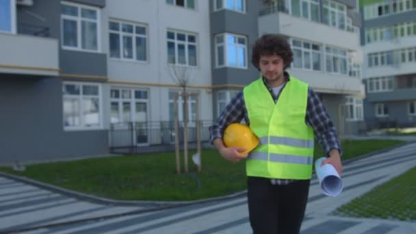 Attractive young architect with black curly hair holding in hand the drawing, going to the camera, putting on head the helmet Outdoor. - Filmmaterial, Video