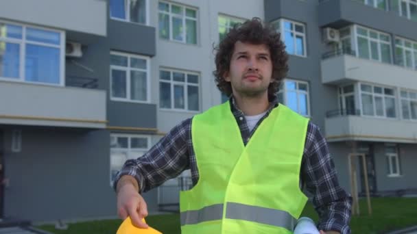 Caucasian young builder with black curly hair holding in hand the drawing, putting on head the helmet, composing the hands. Outdoor. - Footage, Video