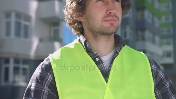 Portrait of young builder with black curly hair in yellow protective helmet looking drawing on the background built house. - Кадры, видео