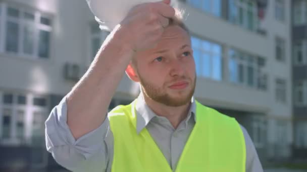 Portrait of handsome builder with rousse hair and beard putting on head white protective helmet on the background built house. - Filmati, video