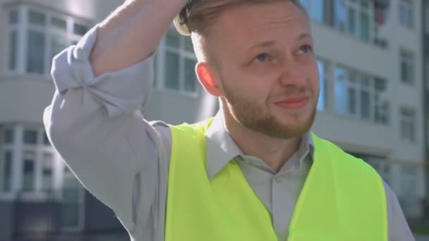 Portrait of attractive builder with rousse hair and beard putting on head white protective helmet and looking drawing on the background built house. - Video