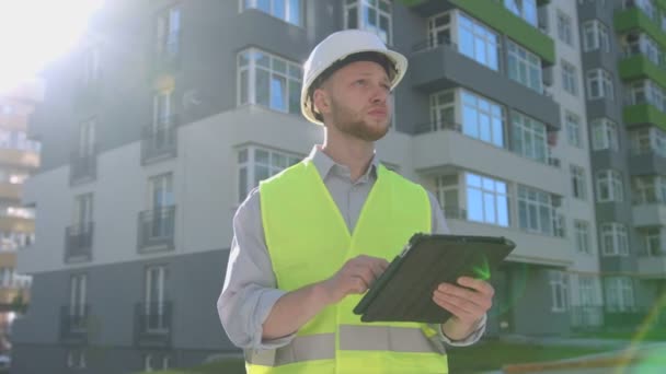 Caucasian handsome builder with white protected helmet and green safety vest using tablet for fixing the stage of construction on the background built house. - Video
