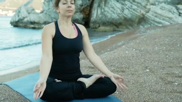Een volwassen vrouw zit in een lotus-positie op het strand in de buurt van stenen stenen. - Video