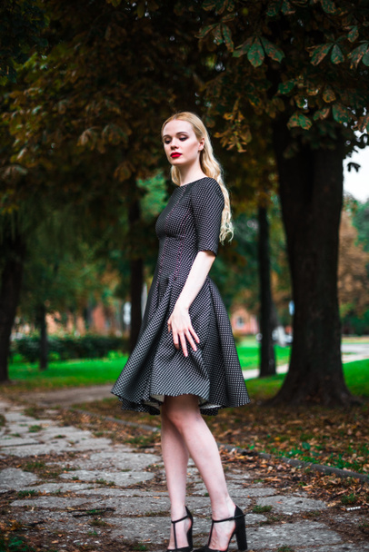 young beautiful blond girl with red lipstick in her big bright eyes and make it in dress posing on the streets , the setting sun trendy clothes - Photo, image